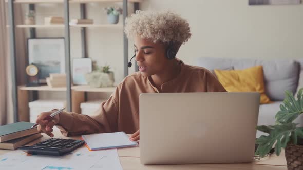 Afro Woman Wears Headset Talking with Customer Calculate Bills Online on Laptop