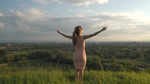 Young woman in pink summer dress standing in green meadow raising her hands up enjoying sunset view.