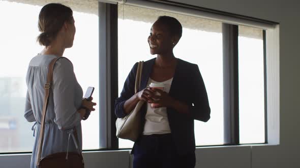 Two diverse female colleagues standing at window, talking and smiling