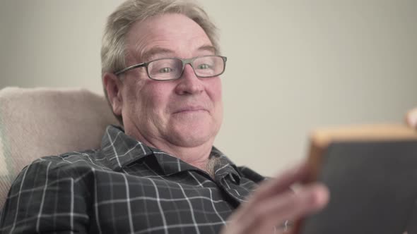 Close-up Portrait of Positive Senior Caucasian Man in Eyeglasses Reading Book and Laughing