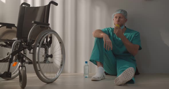 Young Male Nurse or Surgeon in Scrubs Sitting on Hospital Floor and Eating Apple