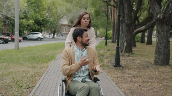 Slow Motion of Caring Young Lady Pushing Man's Wheelchair Talking Smiling Outdoors in Park