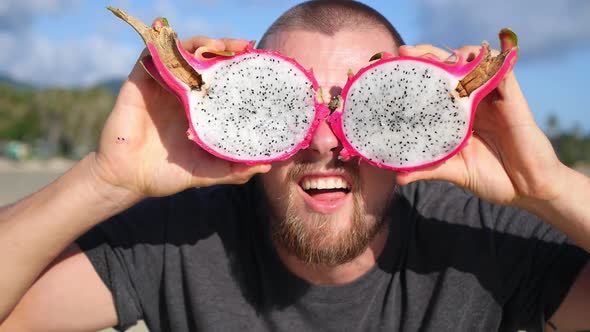 Funny Young Man Holding Up Pink Pitaya Fruit Instead of Eyes At Beach
