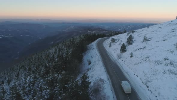 Drone Follows White Truck Trough Snowy Mountain Road Near Frozen Evergreen Forest with Panoramic