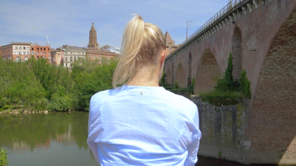 A blonde woman with her hair tied back using her smartphone in front of a river with a bridge on the