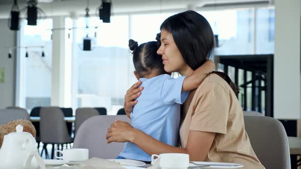 Young Toddler Girl Hugs Mum with Smile Near Table in Cafe