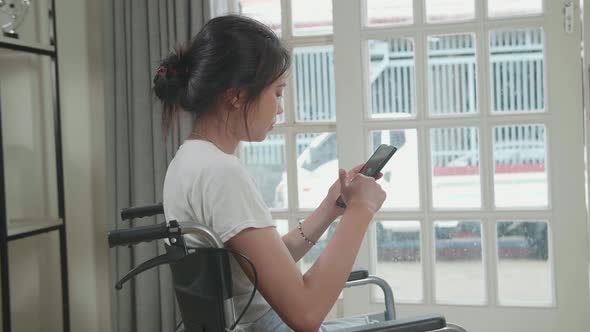 Asian Girl Sitting In A Wheelchair Using Mobile Phone At Home