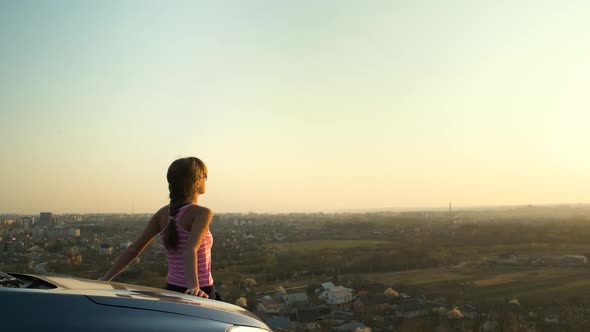 Young woman standing near her car enjoying warm sunset view. Girl traveler leaning on vehicle 