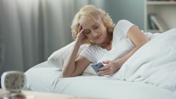 Smiling Mature Woman Lying in Bed and Checking E-Mail on Smartphone, Morning