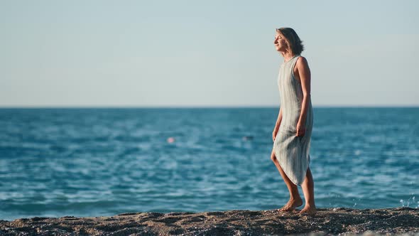 Attractive Tanned Travel Woman Wearing Dress Walking on Beach at Sunset Full Shot