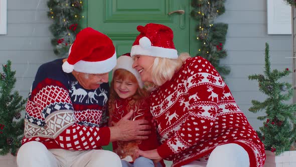 Senior Grandparents with Granddaughter in Santa Claus Hat Celebrating Christmas Near Decorated House