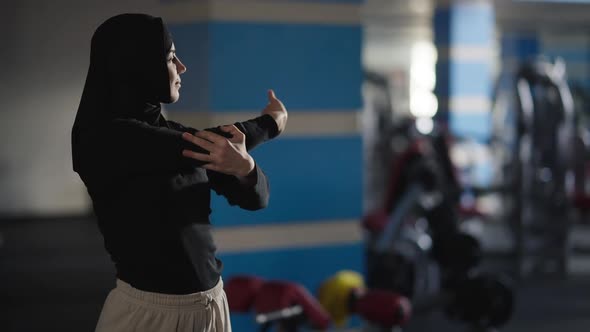 Happy Motivated Woman in Hijab Warming Up Muscles Stretching Hands and Waist Standing Indoors in Gym