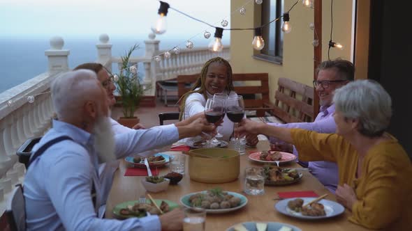 Multiracial senior friends having fun dining together and toasting with red wine on house patio