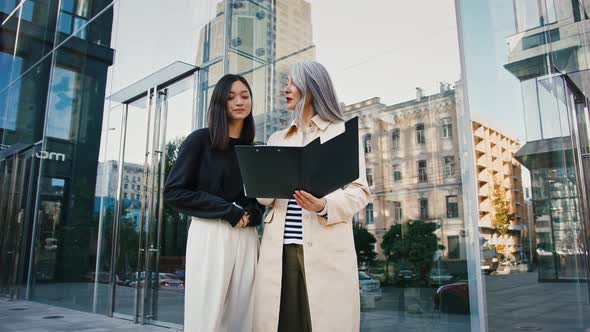 Asian Mature Woman Holding Folder and Explaining Details of New Project to Young Colleague Pointing