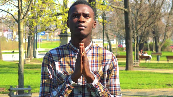 A Young Black Man Prays with Hands Clasped Together in a Park on a Sunny Day