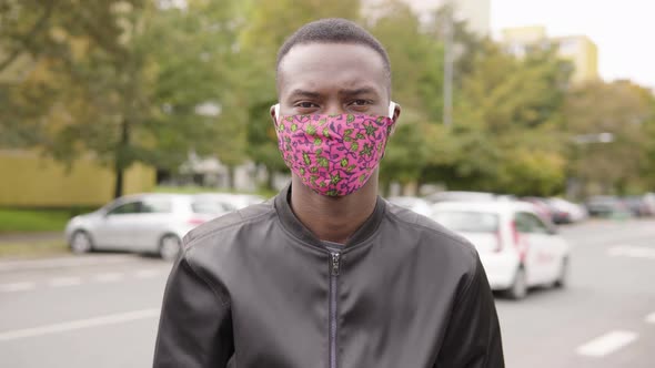 A Young Black Man in a Face Mask Points at the Camera and Nods in a Street in an Urban Area