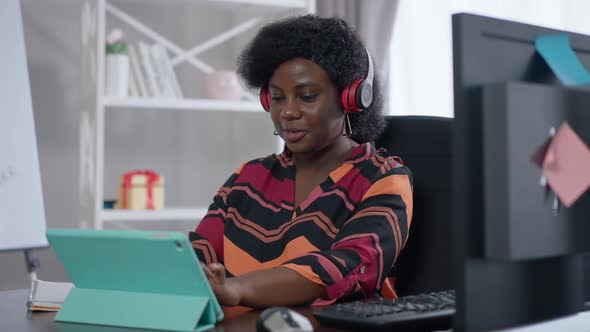 Positive Confident African American Woman in Headphones Using Video Chat on Tablet Sitting in Home