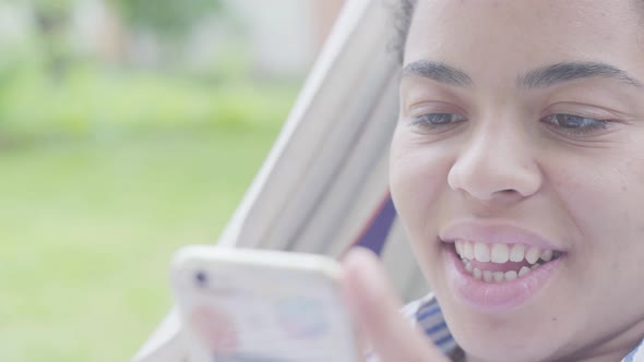 Close-up Face of Happy African American Woman Lying in the Hammock, Relaxing in the Garden, Texting