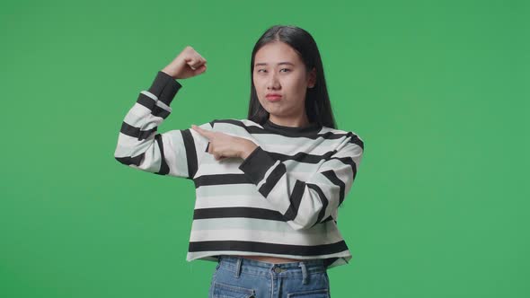 A Smiling Asian Woman Pointing Bicep While Standing In Front Of Green Screen Background