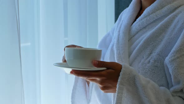 A beautiful young dark-skinned mixed-race woman stands by the window in the hotel and drinks coffee.