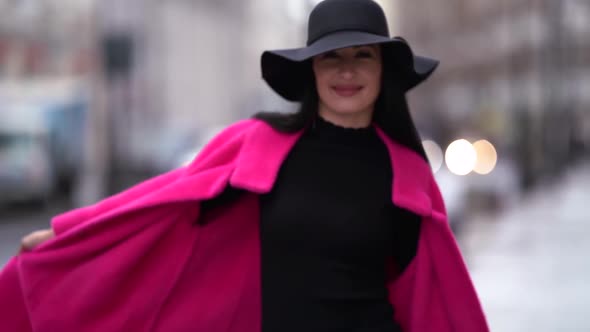 A Stylish Black-haired Woman in a Pink Coat and Black Hat Walks on a Blurry Background of a City