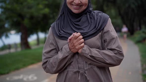 Unrecognizable Smiling Senior Woman in Hijab Putting Hands Together Standing on Park Alley