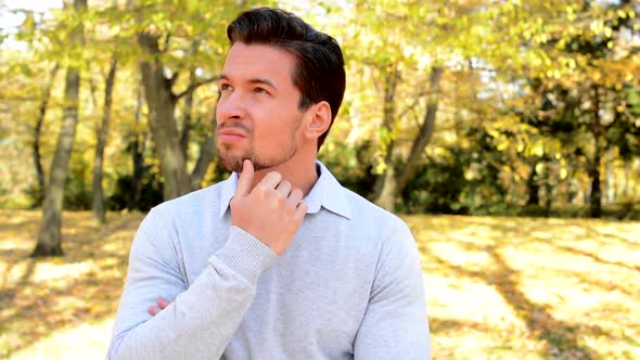 Young Handsome Man Stands in the Woods, Strokes His Chin, Looks Worried and Thinks About Something