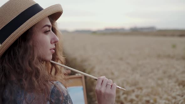 Pretty Paintress in Hat and Dress Posing with Brush Among Ripe Wheat Field