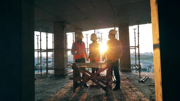Workers Talk in Unfinished Building on a Sunset Background. Multiethnic Engineers, Architects alt