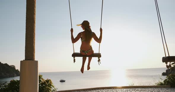 Young Woman in Hat Swaying on Rope Swing at Sunset on the Beach with Seaview