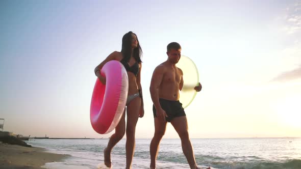 Happy Young Couple Walking on the Beach with Rubber Rings During Sunset Slow Motion