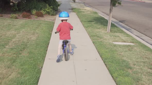 Follow shot of a young boy riding a blue balance bicycle along a sidewalk in a residential neighborh