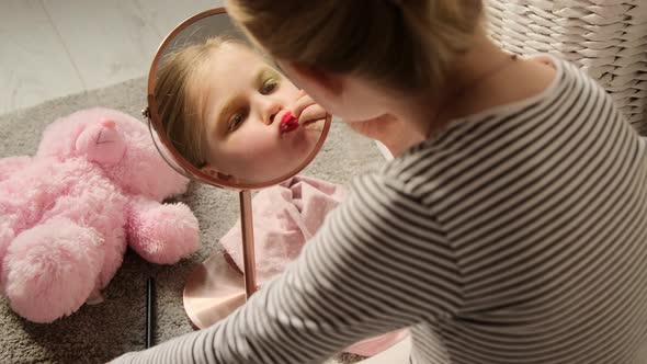 Little child girl paints her lips with lipstick, plays makes learn makeup in front of the mirror