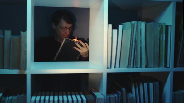 Man in Glasses with Candle Reading Book Standing Behind Bookshelf