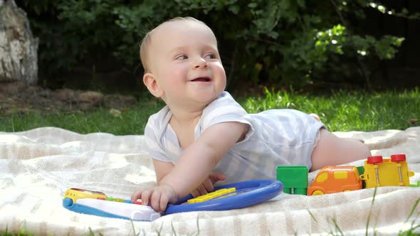 Cute Smiling Baby Boy Lying on Grass at Garden and Playing with Colorful Toys