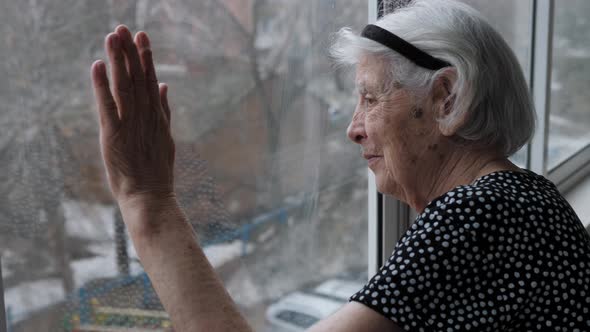 Old Elderly Grandmother Woman Looks Calm Out Of Window With Hand On The Glass