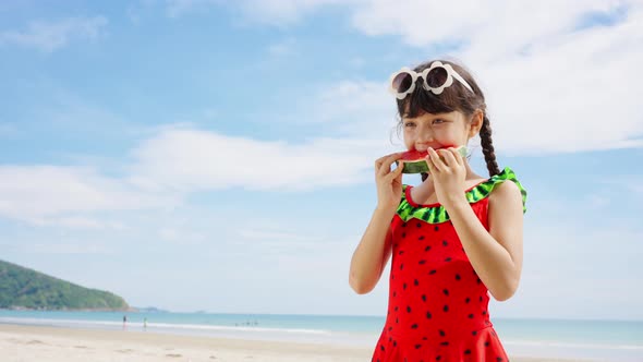 4K Little Asian girl in swimwear eating watermelon while playing on the beach in summer sunny day.