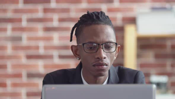 Serious office black man in glasses and a jacket sits at desk, working on a laptop.