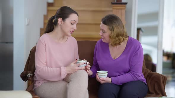 Relaxed Cheerful Adult Mother and Young Daughter Talking Smiling Sitting on Comfortable Couch with