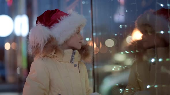 Little Girl in Santa Hat with Gifts Box Looking and Dreaming in Christmas Window Shopping on