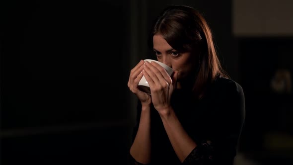 Young Naive Woman with Big Cup of Tea or Coffee Is Sitting in Dark Studio, Interview Concept