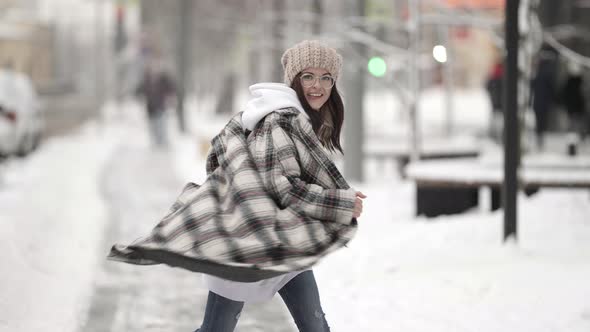 A Darkhaired Woman in Round Glasses Jumps and Spins Merrily on a City Street on a Winter Evening