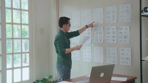 Young Creative Asian Designer Looks At The Storyboard Sketches Covering His Wall