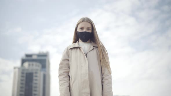 Middle Shot Portrait of Young Woman in Covid Face Mask Standing in City at the Background of Cloudy