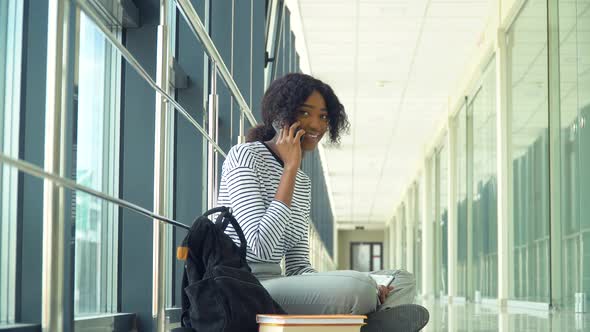 African American Woman Student Sitting on the Floor Talking on the Phone in the University Coridor