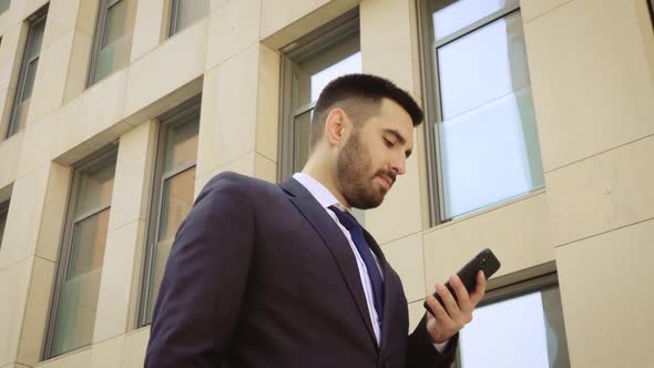 Close Up View of Smiling Businessman Dressed in Shirt Using Smartphone While Walks on the Background