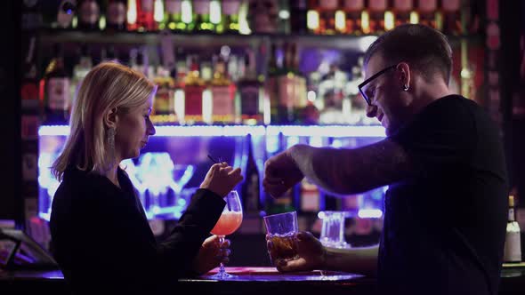 A Beautiful Couple Drink Cocktails at the Bar Counter. Man in Glasses and Blond Woman Talking