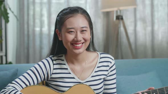 Close Up Of Asian Woman Looking And Smiling To Camera While Playing A Guitar At Home