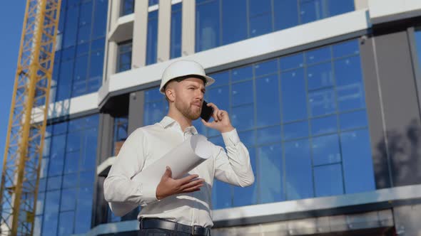 An Engineer in a White Shirt and Helmet Works on the Construction of a Modern Glass Building