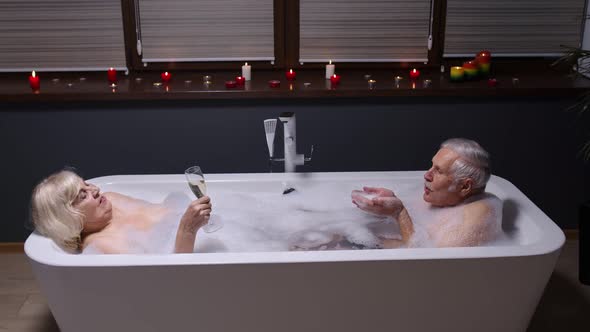 Senior Couple Grandfather and Grandmother Is Taking Foamy Bath, Drinking Champagne in Bathroom